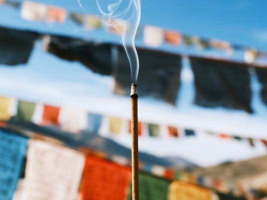 A Tibetan incense stick releasing smoke against a backdrop of colorful prayer flags in the mountains.