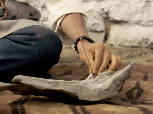 Close-up of a hand gently placing a thick Tibetan incense stick into a driftwood holder, set on a woven mat with traditional patterns. The person wears a bead bracelet and sits cross-legged in a rustic setting.