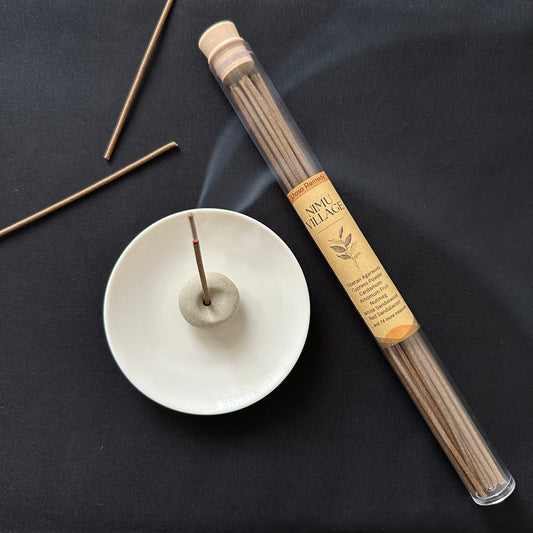 Nimu Village Tibetan incense sticks in a glass tube on a dark background with a white plate and a ceramic holder holding a burning incense stick.
