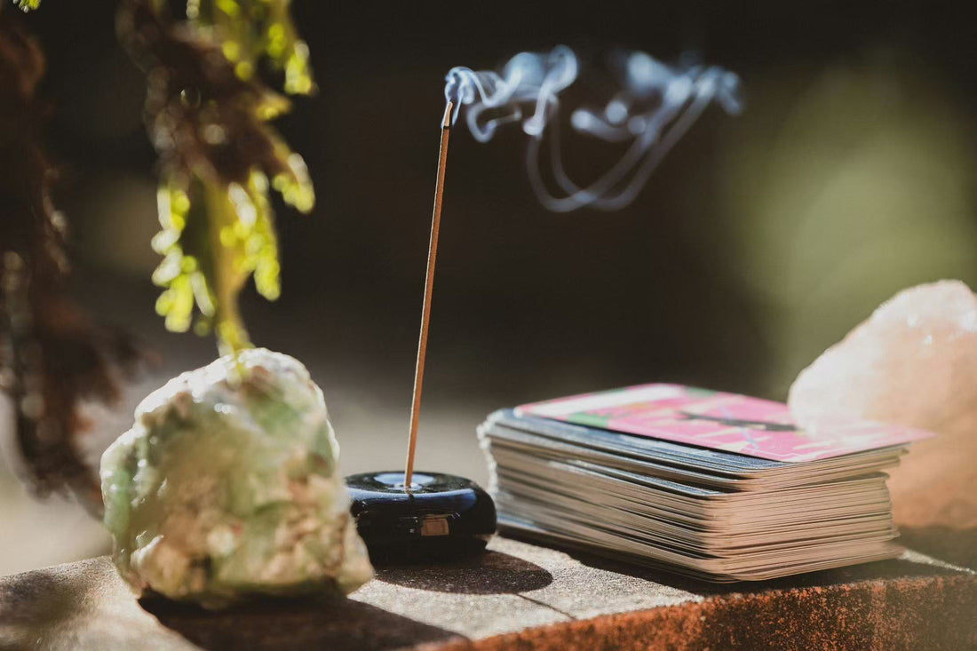 A lit incense stick releasing delicate wisps of smoke, placed in a black holder alongside a green crystal, a stack of tarot cards, and a rose quartz crystal, bathed in warm sunlight.