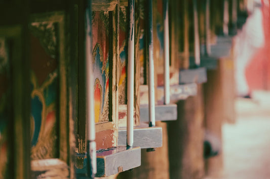 A close-up of traditional Tibetan prayer wheels, ornately painted with vibrant patterns in red, blue, and gold tones, mounted on a wooden structure in a monastery or sacred site.