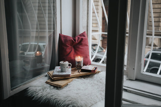 A cozy window nook featuring a lit candle, books, and a warm drink, illustrating why some people prefer candles over incense for relaxation rituals. Highlights the candles vs incense preference in spiritual and daily routines.