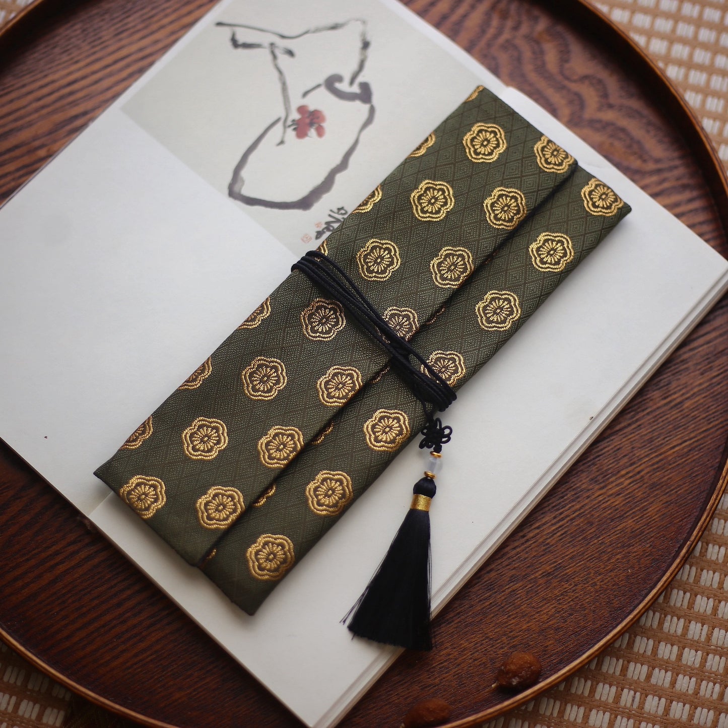 Olive-green silk incense carry pouch with gold floral embroidery and a black tassel, placed on an open art book atop a wooden tray