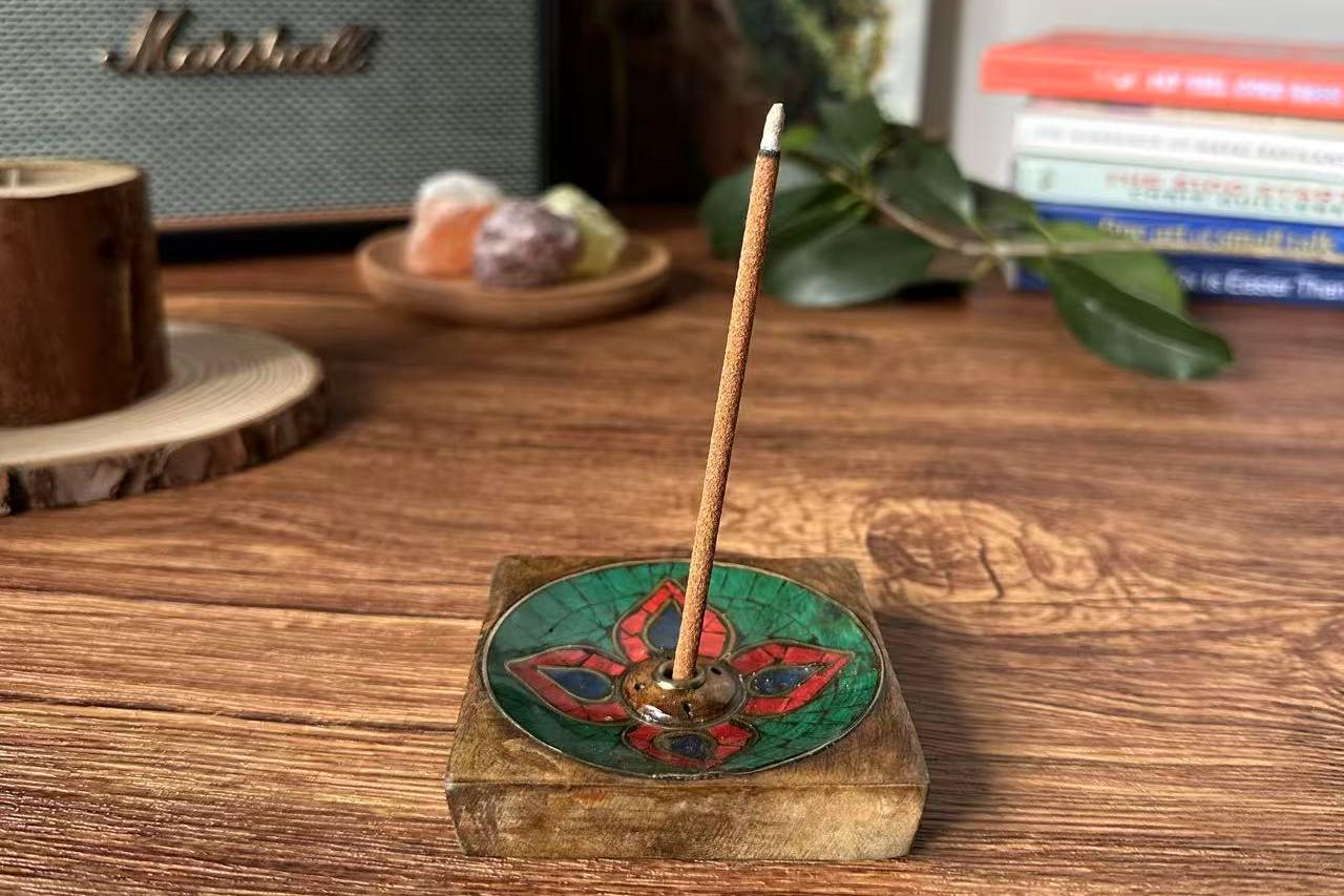 A lit Tibetan incense stick burning in a handcrafted wooden and mosaic incense holder with red and green floral patterns. The scene is set on a wooden table with crystals, books, and a speaker in the background, creating a cozy and mindful atmosphere.