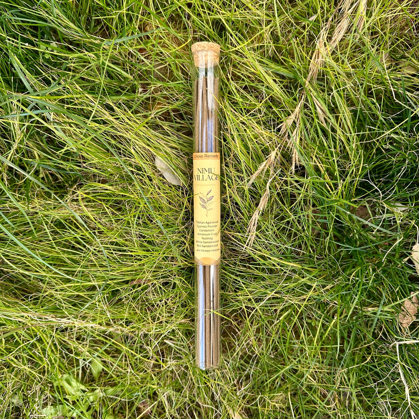 A glass tube of Lhasa Remedy Nimu Village incense sticks with a cork lid, resting on a bed of natural green grass.