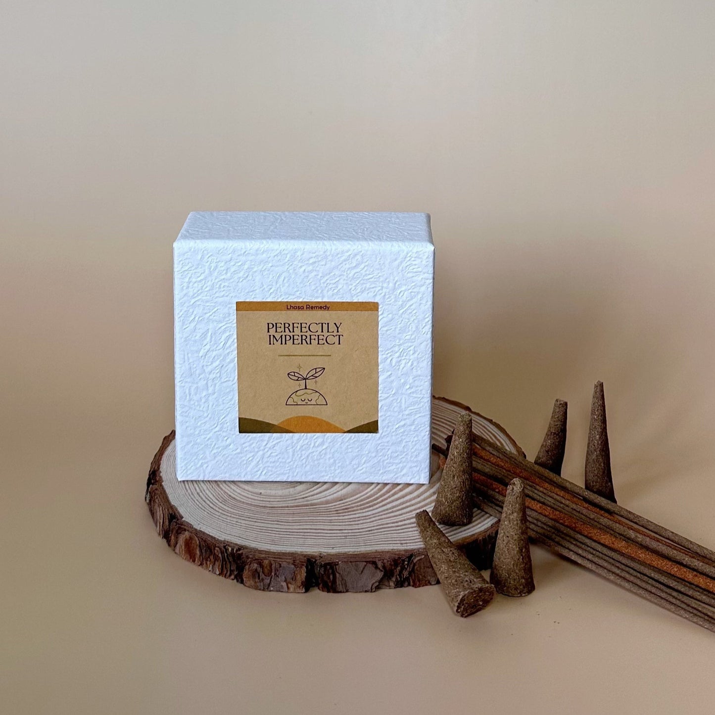 A white textured box labeled "Perfectly Imperfect" from Lhasa Remedy sits atop a wooden slab, accompanied by Tibetan incense sticks and cones, against a neutral beige background, creating a minimal and earthy aesthetic.