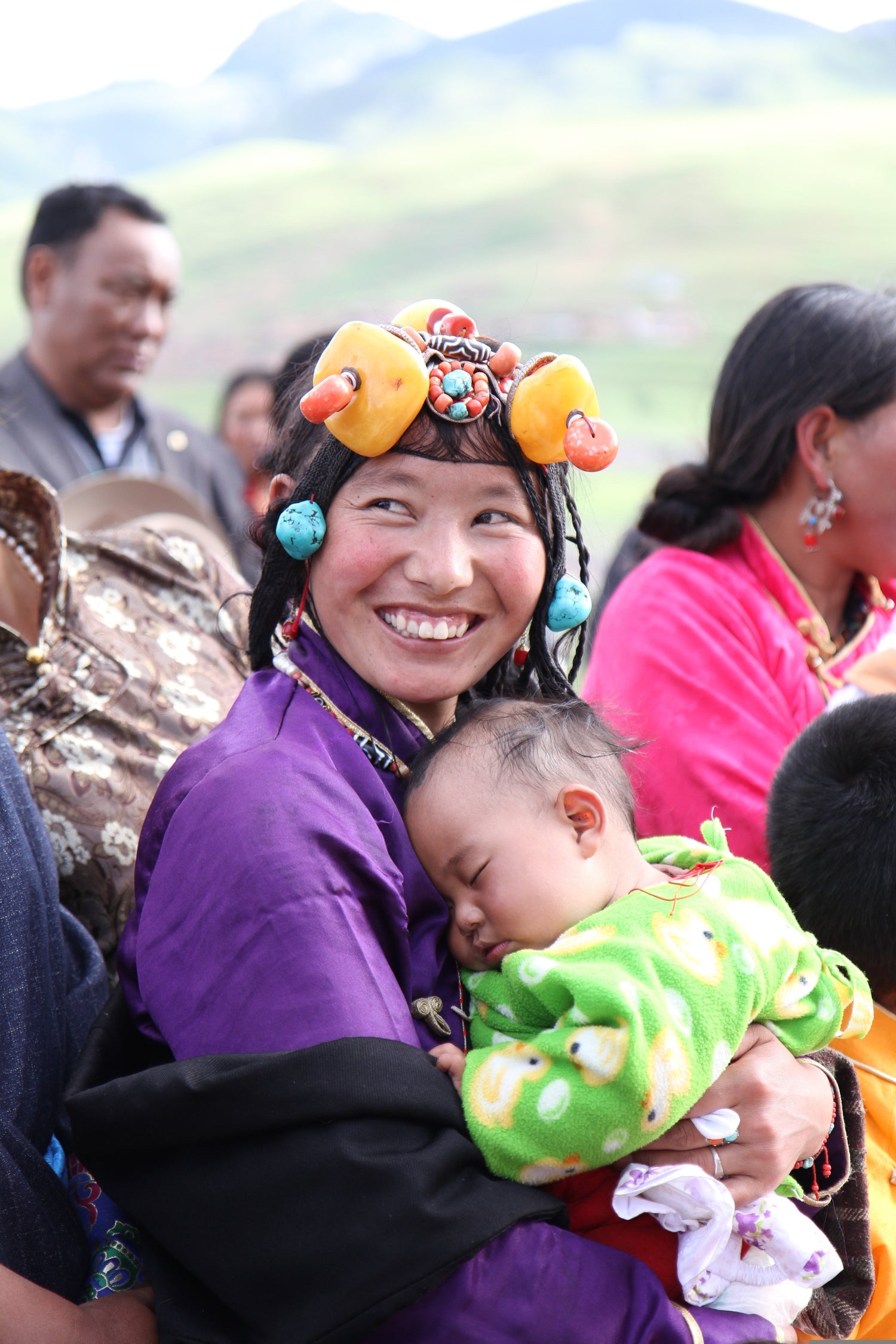 Tibetan mother in traditional attire smiling while holding her sleeping baby during a cultural gathering.