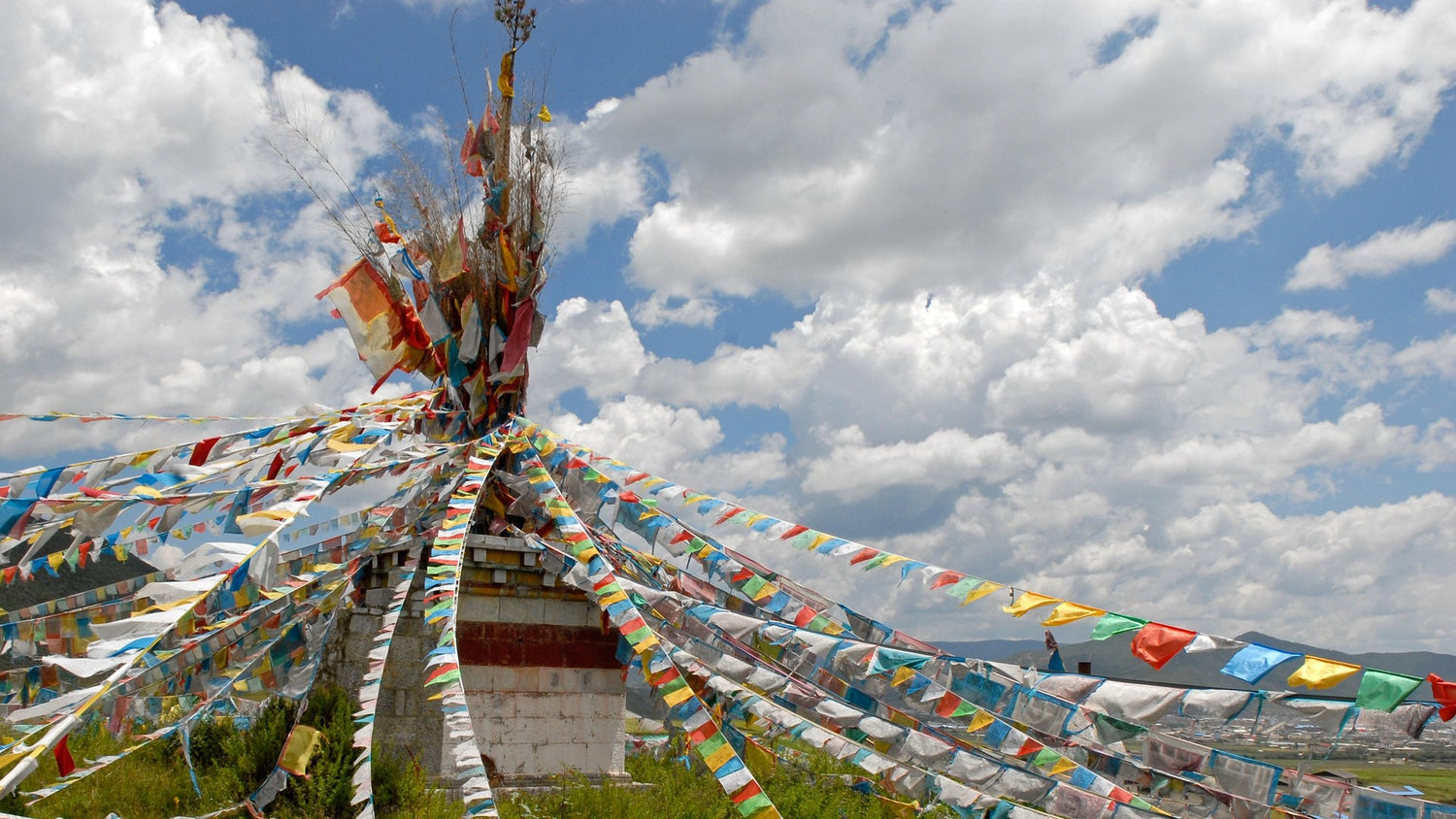 Long strands of colorful Tibetan prayer flags radiating from a central pole, set against a dramatic cloudy sky and distant mountain village in the background.
