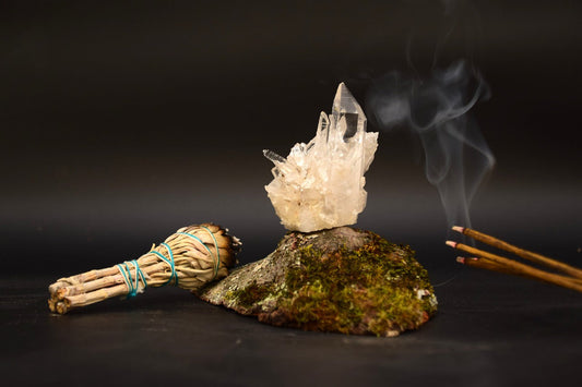 A natural rock displaying a crystal on its surface, with a few burning incense stick and a bundle of herbs placed next to it against a black background, illustrating a serene outdoor scene.
