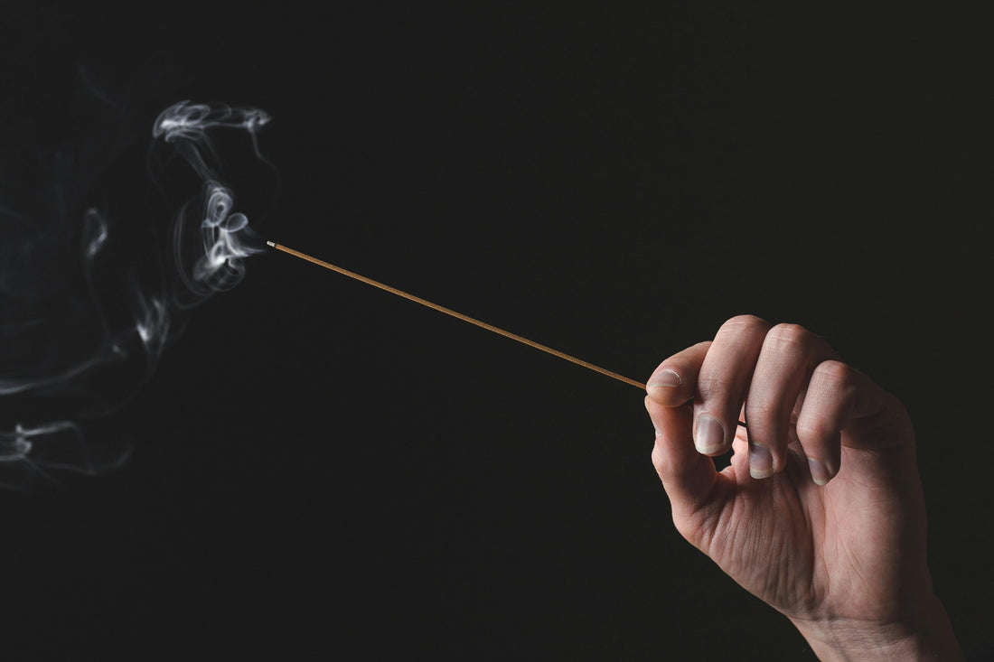 An individual gently holds an incense stick with one hand, releasing fragrant smoke that swirls into the air  against the black background.