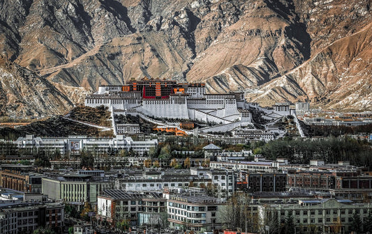 Majestic Potala Palace in Tibet, an iconic symbol of Tibetan culture, surrounded by houses and mountains, rises beautifully on the top of a hill against the mountainous backdrop and a clear blue sky.