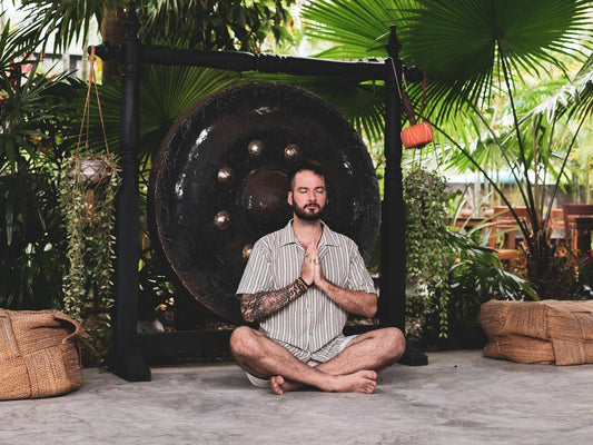 Man sitting cross-legged with eyes closed and hands in prayer position, meditating in front of a large gong surrounded by tropical plants in an outdoor retreat setting.