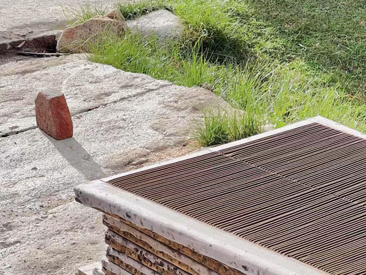 Stacked trays of freshly made Tibetan incense sticks drying under the sun on stone ground near grass, using traditional mesh screens for airflow.