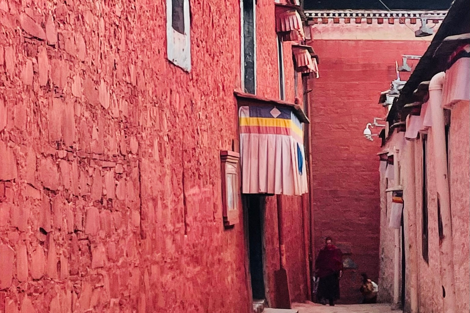 A Tibetan monk walks down a narrow alley in a temple, flanked by a vibrant red wall.
