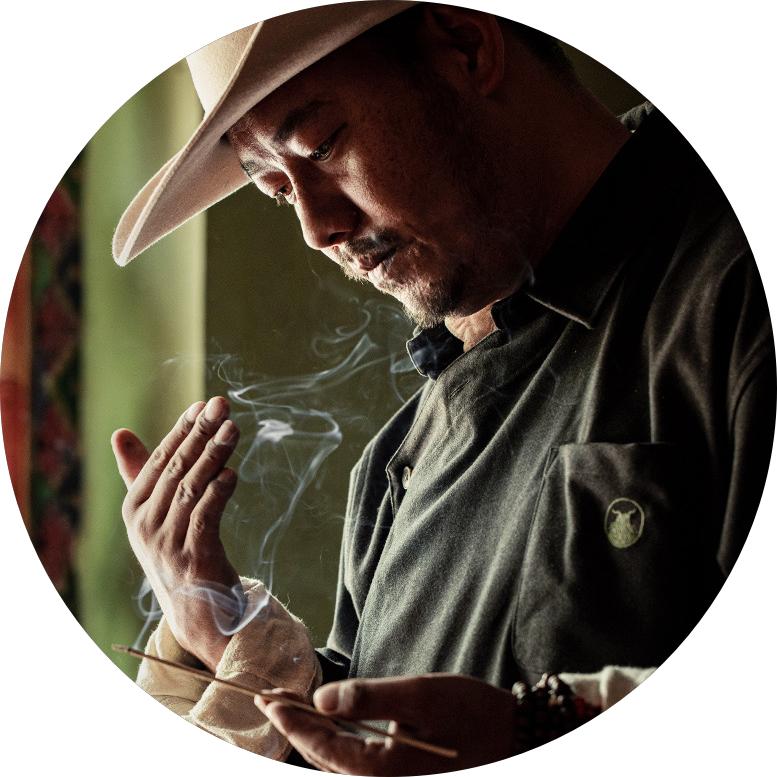 A man wearing a hat and traditional Tibetan clothes is holding a burning incense stick with his left hand and enjoying the fragrant smoke of the incense.