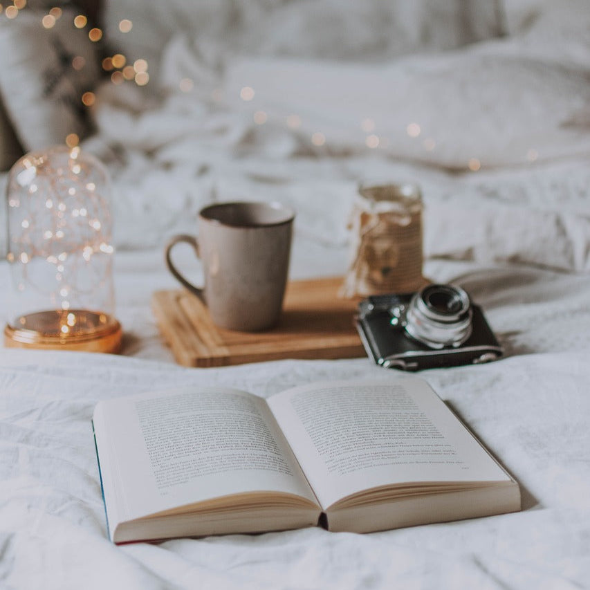 A book rests on a bed, illuminated by a lamp, with a cup of coffee nearby, creating a warm reading atmosphere.