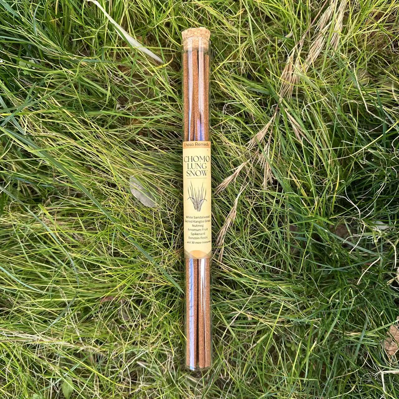 A glass tube placed in vividly green grass, with Lhasa Remedy's Chomolung Snow Tibetan incense sticks inside and a yellow product label on the tube.