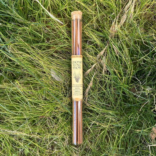 A glass tube placed in vividly green grass, with Lhasa Remedy's Chomolung Snow Tibetan incense sticks inside and a yellow product label on the tube.