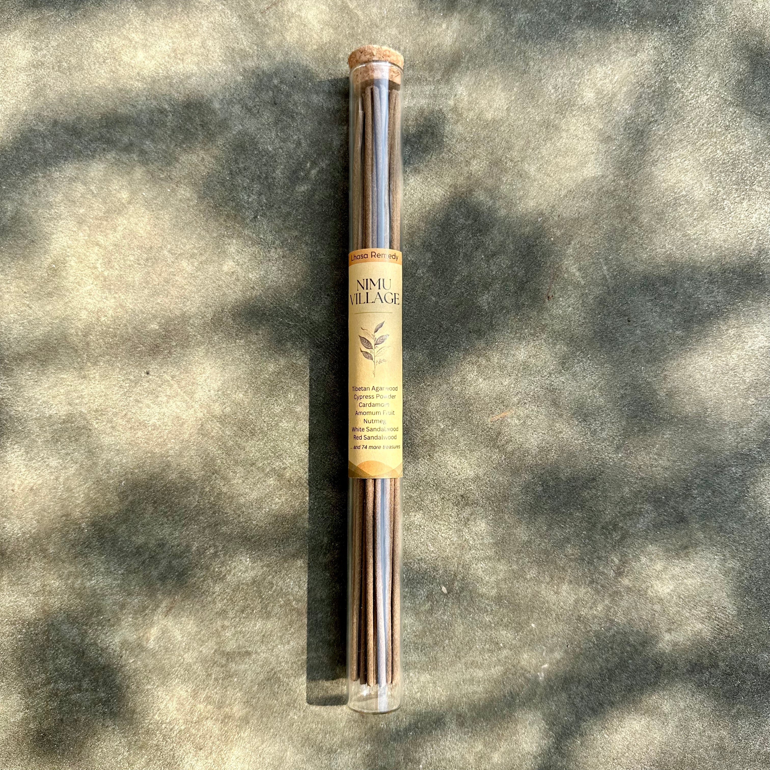 A glass tube positioned on the stone ground, with Tibetan incense sticks in it and a product label of "Nimu Village" sticking onto it, covering by sunshine and shadows.
