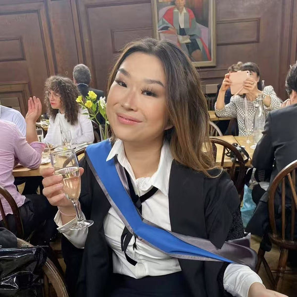 Co-founder of Lhasa Remedy LeLe celebrating her graduation with a toast, dressed in academic robes and smiling confidently in a lively dining hall.