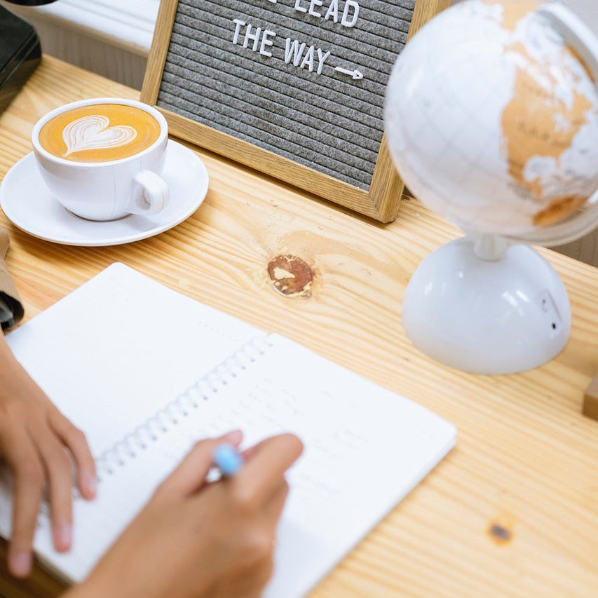 A person writes in a notebook with a pen, surrounded by a cup of coffee, a globe, and a board, symbolizing love's guidance.