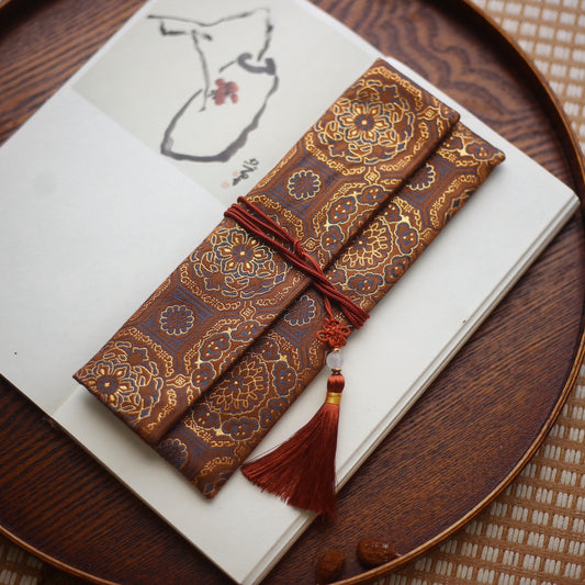 Bronze silk incense carry pouch with golden lotus embroidery and a deep red tassel, placed on a wooden tray with an art book