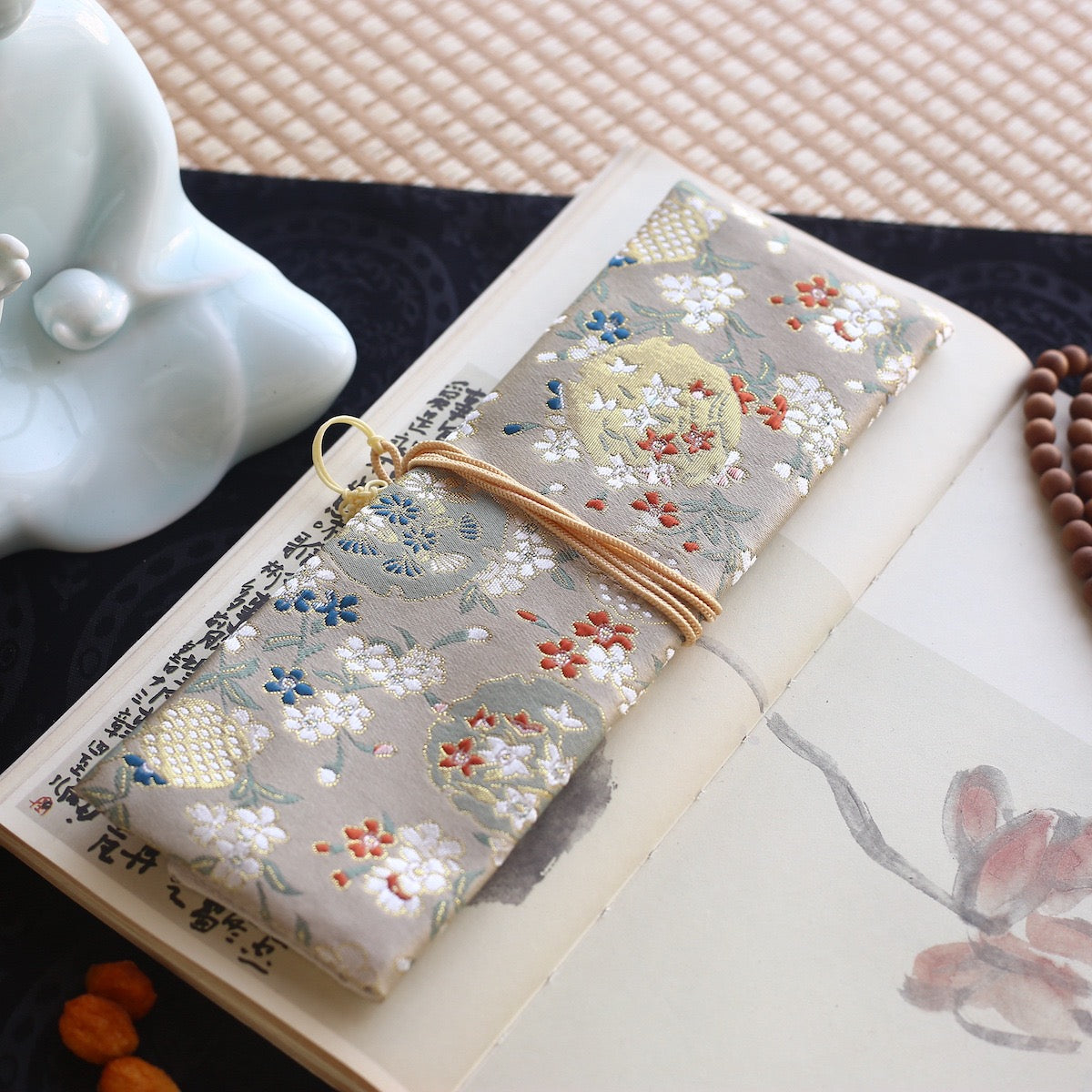 Ivory silk incense carry pouch with colorful floral embroidery, tied with a golden cord and displayed on an open book