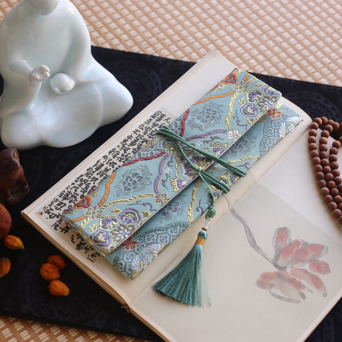 Soft jade-blue silk incense pouch with ornate palace-style patterns and a teal tassel, styled atop a traditional book