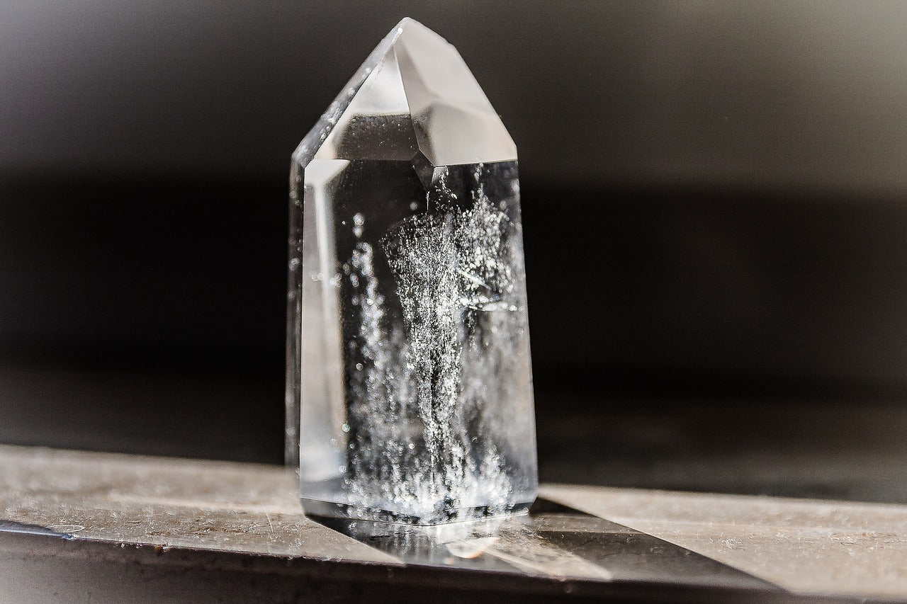 A clear quartz crystal standing upright on a reflective surface, with natural inclusions and internal fractures visible inside its transparent structure, illuminated by soft light.