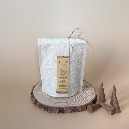 A pack of Lhasa Remedy’s Lucky Zaki Tibetan incense cones in eco-friendly handmade paper packaging, tied with twine, placed on a wooden slice. Several incense cones are arranged beside it against a neutral beige background.