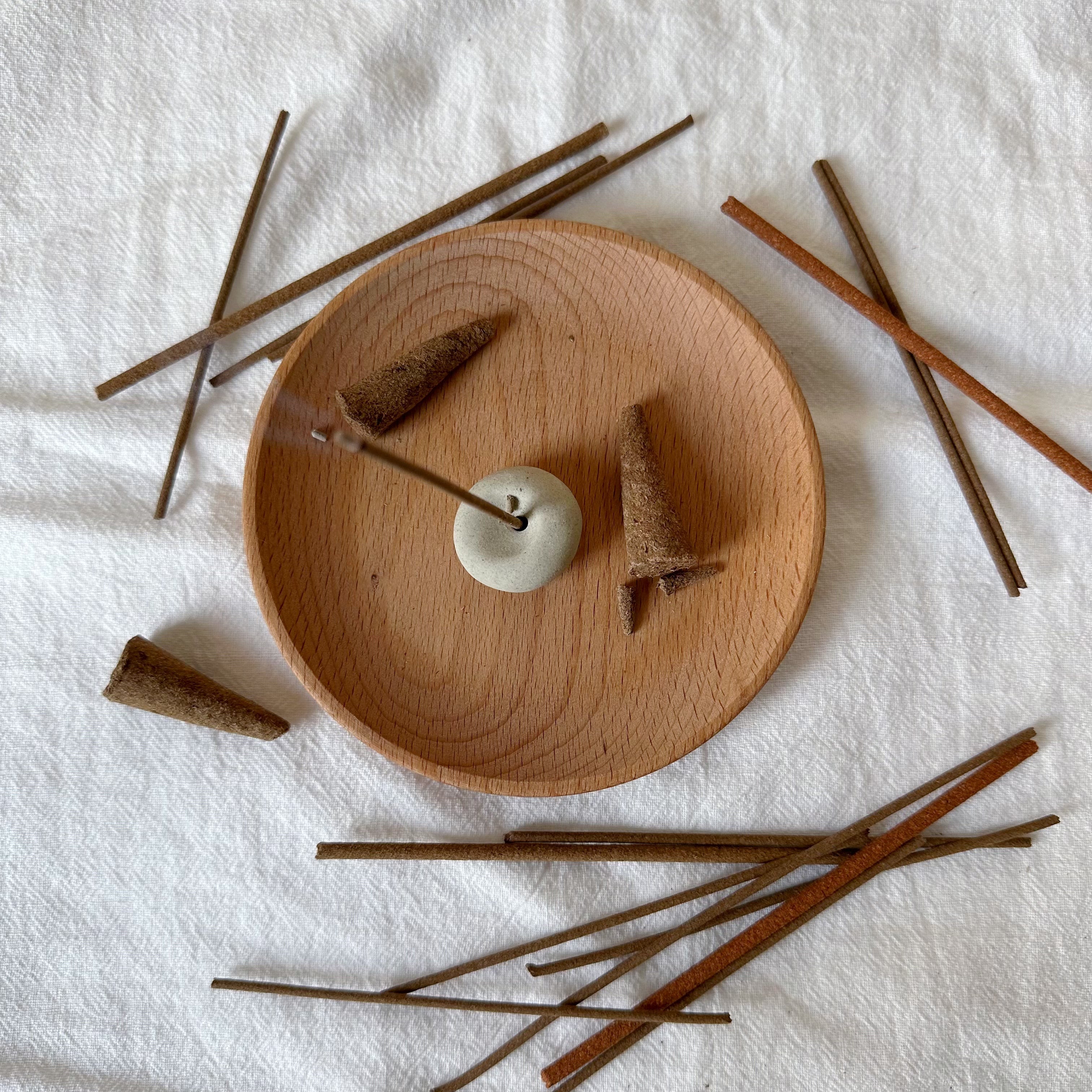 A collection of Tibetan incense sticks and cones arranged on a round wooden tray with a minimalist ceramic holder, placed on a soft white fabric background, creating a serene and natural aesthetic.